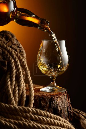 Foto de El brandy se vierte de una botella en un vaso. Vaso con whisky, coñac o brandy en un tocón viejo. - Imagen libre de derechos
