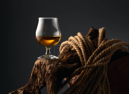 Téléchargez les photos : Brandy snifter et corde sur un vieux chevalet en bois. Verre avec whisky, cognac ou brandy sur fond sombre. - en image libre de droit