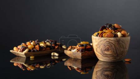 Téléchargez les photos : Mélange de diverses noix et raisins secs dans un plat en bois sur un fond réfléchissant noir. - en image libre de droit