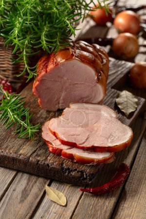 Foto de Sabroso jamón de cerdo ahumado con romero, cebolla, ajo, pimiento rojo y hojas de laurel. Carne ahumada en una mesa de madera. - Imagen libre de derechos