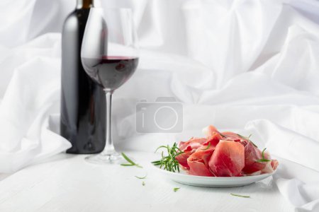 Téléchargez les photos : Prosciutto au romarin et vin rouge sur une table en bois blanc. Snack italien traditionnel. - en image libre de droit