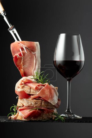 Photo for Ciabatta with prosciutto, rosemary and glass of red wine on a black background. Traditional Italian snack. - Royalty Free Image