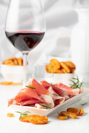 Photo for Prosciutto, spicy bruschetta crackers and glass of red wine on a white wooden table. Traditional Italian kitchen. - Royalty Free Image