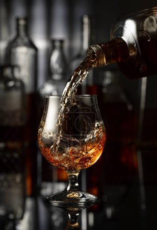 Téléchargez les photos : Verser le brandy d'une bouteille dans un verre. Un soupçon de brandy sur une table noire dans un bar. Sur le fond bouteilles avec diverses boissons alcoolisées. - en image libre de droit