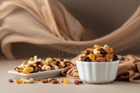 Photo for The mix of various nuts and raisins in a white bowl on a beige background. In the background there is a beige curtain fluttering in the wind. Copy space. - Royalty Free Image