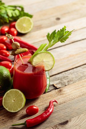Foto de Cóctel Bloody Mary decorado con pepinillo, olivo, lima y apio. Bebida de tomate con ingredientes en una vieja mesa de madera. - Imagen libre de derechos