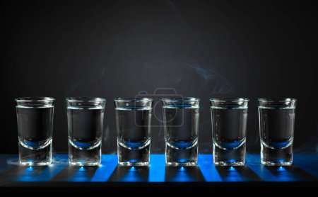 Foto de Las tomas alcohólicas del vodka o la bebida fuerte en los vasos pequeños sobre el fondo negro. Luz de fondo azul. Copiar espacio. - Imagen libre de derechos