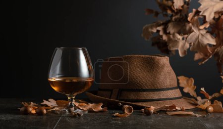 Téléchargez les photos : Snifter de brandy sur une table en pierre avec des feuilles de chêne séchées et un chapeau d'homme. - en image libre de droit