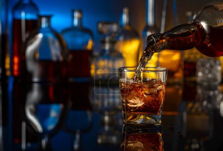 Foto de El whisky se vierte de una botella en un vaso con hielo. Un vaso de whisky sobre un fondo negro reflectante. En las botellas de fondo con varias bebidas alcohólicas. - Imagen libre de derechos