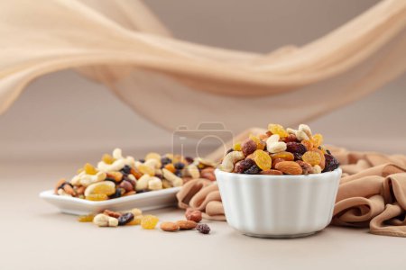 Téléchargez les photos : Le mélange de diverses noix et raisins secs dans un bol blanc sur un fond beige. En arrière-plan, il y a un rideau beige flottant dans le vent. Espace de copie. - en image libre de droit