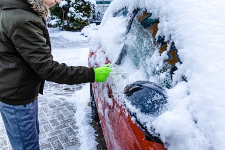 Téléchargez les photos : Teenager  cleans car after a snowfall, removing snow and scraping ice - en image libre de droit