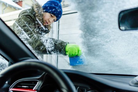 Foto de Teenager  cleans car after a snowfall, removing snow and scraping ice - Imagen libre de derechos