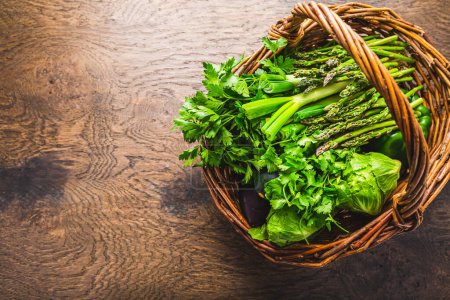 Foto de Verduras y hierbas verdes frescas en canasta de mimbre, espárragos, lechuga, calabacín con perejil y cilantro sobre mesa de madera - Imagen libre de derechos