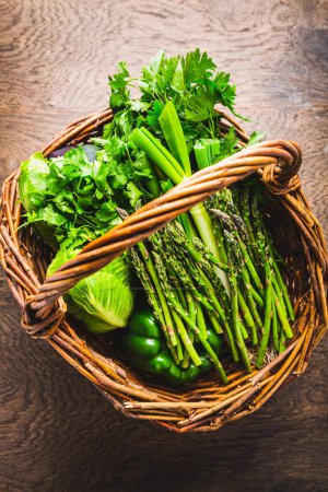 Photo for Fresh green vegetables and herbs in wicker basket, asparagus, lettuce, zucchini with parsley and cilantro  on wooden table - Royalty Free Image