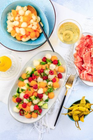 Foto de Comida ligera de verano. Ensalada de pepino y melón con frambuesas y jamón y pimientos picantes en escabeche - Imagen libre de derechos