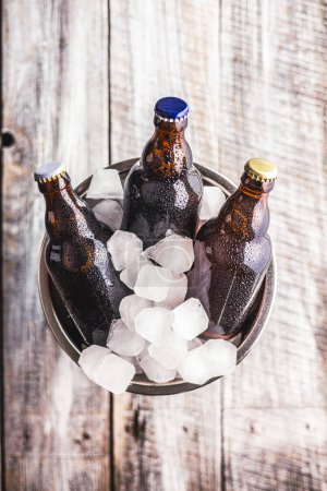 Un seau en métal est rempli de glace, et trois bouteilles de bière en verre sont placées à l'intérieur, refroidissant.