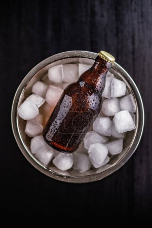 A metal bucket is filled with ice, and one  glass bottles of beer is placed inside it, chilling.