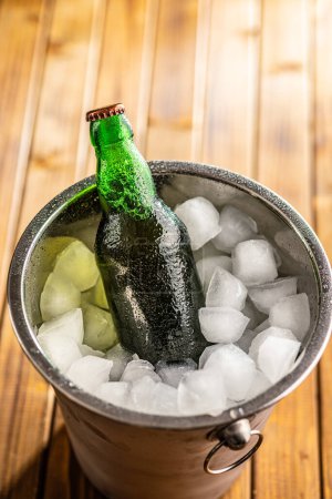 Un seau en métal est rempli de glace, et une bouteille de bière en verre est placée à l'intérieur, refroidissant.