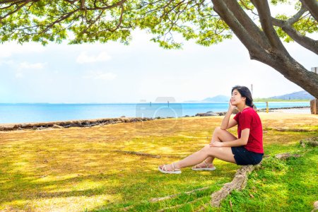 Téléchargez les photos : Jeune femme reposant à l'ombre d'un grand arbre à baldaquin à Hawaï, avec l'horizon océanique en arrière-plan - en image libre de droit
