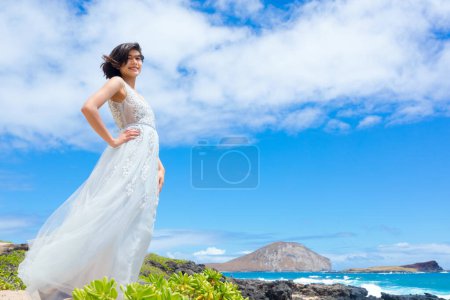Foto de Sonriente adolescente en vestido blanco de pie junto a rocas de lava rocosa en la playa hawaiana por el océano - Imagen libre de derechos