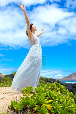 Foto de Chica adolescente en vestido blanco levantando brazos en alabanza al aire libre en la costa hawaiana - Imagen libre de derechos