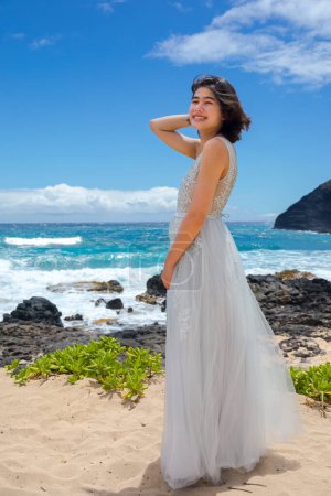 Foto de Sonriente adolescente en vestido blanco de pie junto a rocas de lava rocosa en la playa hawaiana por el océano - Imagen libre de derechos