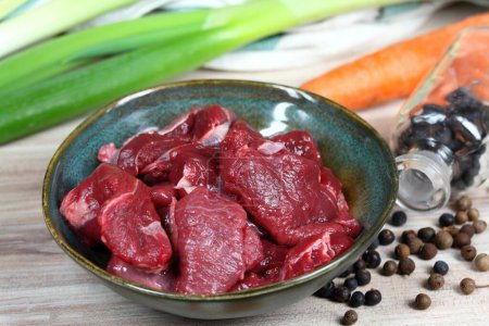 Rohes Hirschfleisch für Hirschragout oder Gulasch. Schüssel mit Hirschfleisch, frischem Gemüse, Wacholderbeeren und Piment zum Kochen.