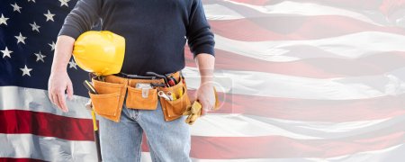 Photo for Male Contractor Wearing Tool Belt and Holding Blank Yellow Hardhat and Gloves Over Waving American Flag Background Banner. - Royalty Free Image