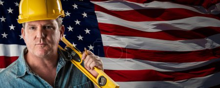 Photo for Male Contractor Wearing Blank Yellow Hardhat Over Waving American Flag Background Banner. - Royalty Free Image