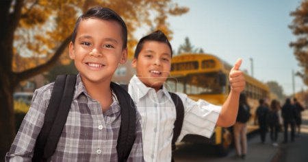 Téléchargez les photos : Deux jeunes garçons hispaniques heureux portant des sacs à dos donnent un pouce sur le campus près d'un autobus scolaire - en image libre de droit