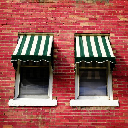 Two windows in brick wall on building apartment with green striped awnings or window shades