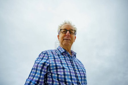 Téléchargez les photos : Portrait of a senior man in a blue shirt and glasses on a background of a cloudy sky. - en image libre de droit