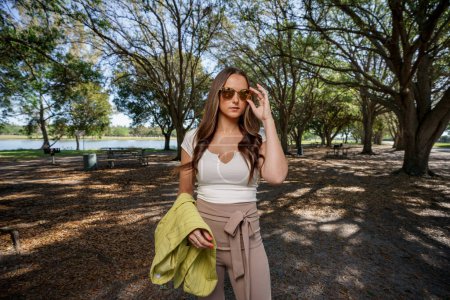 Téléchargez les photos : Photo éclair d'une femme ajustant ses lunettes de soleil posant dans un parc naturel - en image libre de droit
