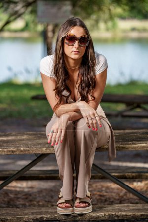 Photo for Photo of a young woman wearing sunglasses sitting on a picnic table in the park - Royalty Free Image