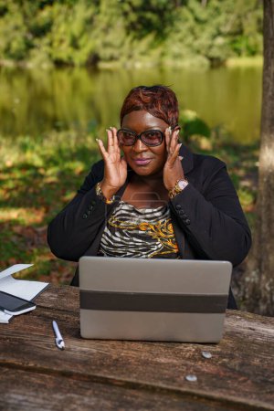 Photo for Businesswoman putting on her glasses. She is sitting on a park bench while at work - Royalty Free Image