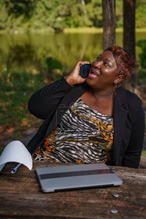 Foto de Exitosa mujer de negocios negra en una llamada de negocios mientras trabaja remotamente en el parque - Imagen libre de derechos