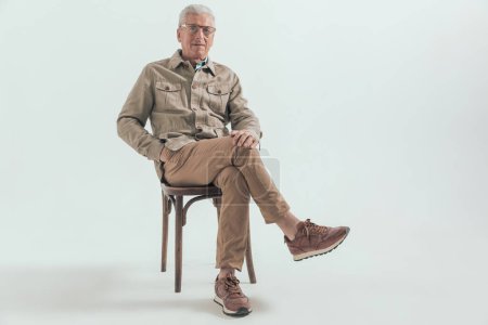 Téléchargez les photos : Vieil homme décontracté se relaxant sur une chaise en bois, croisant les jambes et tenant une main dans la poche - en image libre de droit