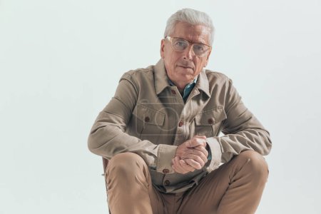 Foto de Viejo hombre casual frotando las palmas y sentado en una posición relajada, con gafas graduadas sobre fondo gris - Imagen libre de derechos