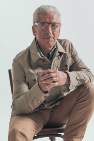 Foto de Modelo de moda vieja sentado en una silla, frotando sus palmas, mirando hacia otro lado y con gafas en el fondo gris del estudio - Imagen libre de derechos
