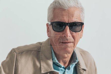 Foto de Primer plano de un viejo hombre de negocios con gafas de sol y una chaqueta crema contra fondo gris estudio - Imagen libre de derechos