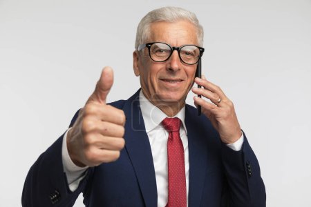 Photo for Businessman giving a ok sign and showing that the conversation on the phone went ok against grey studio background - Royalty Free Image