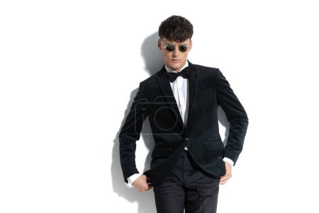 Photo for Dramatic businessman looking down, taking his hands out of his pockets and wearing sunglasses and a black tuxedo against white studio background - Royalty Free Image