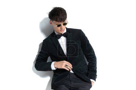 Photo for Dramatic businessman is looking down, putting one hand in pocket, closing his jacket, and wearing a ring and sunglasses on white studio background - Royalty Free Image