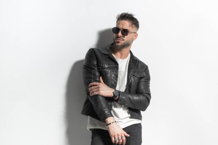 sexy man with wet hair wearing sunglasses and looking away while posing with arms in a fashion way in front of grey background