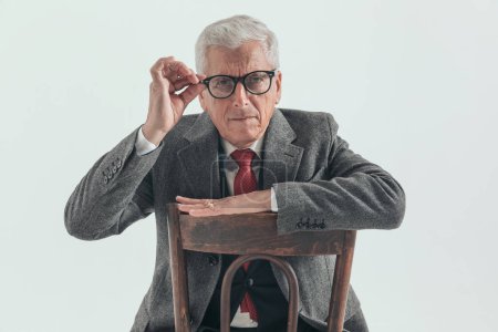 Photo for Curious old man with grizzled hair adjusting glasses and trying to see while sitting and posing with arm on chair back in front of grey background - Royalty Free Image