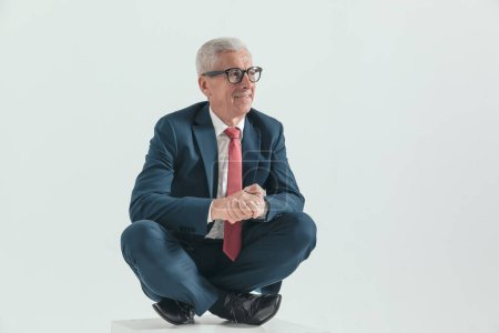 Téléchargez les photos : Homme d'affaires dans la soixantaine avec les cheveux gris regardant vers le côté, tenant la main et assis avec les jambes croisées en dessous devant fond gris - en image libre de droit