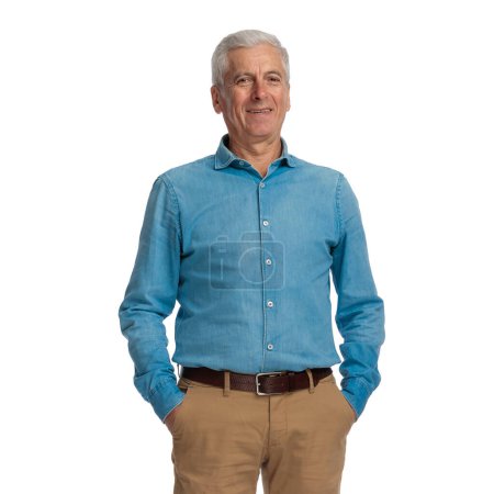 excited old guy with grey hair wearing blue denim shirt and chino pants smiling and posing with hands in pockets in front of white background in studio