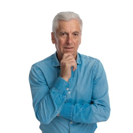 pensive old man in denim shirt folding arms, touching chin, thinking and posing in front of white background in studio