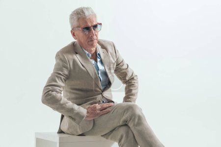 Téléchargez les photos : Homme de mode cool avec les jambes croisées de cheveux gris, regardant loin et posant d'une manière confiante sur fond gris, assis dans le studio - en image libre de droit
