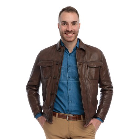 Foto de Portrait of happy casual guy in denim shirt with leather jacket smiling and posing with hands in pockets in front of white background in studio - Imagen libre de derechos